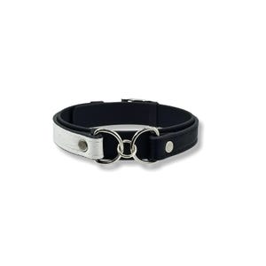 Könnte beinhalten: Ein zweifarbiges Halsband mit einem weißen, ledertexturierten Segment auf der linken Seite und einem schwarzen, ledertexturierten Segment auf der rechten Seite. Beide Seiten sind durch zwei silberfarbene Metallringe und einen kleinen Ring verbunden.
