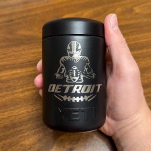 K&ouml;nnte beinhalten: Schwarzer isolierter Becher mit einer silbernen Grafik eines Footballspielers, der einen Football h&auml;lt, dem Wort "DETROIT" und der Marke "YETI". Der Becher ist zylindrisch mit einem schwarzen Deckel.