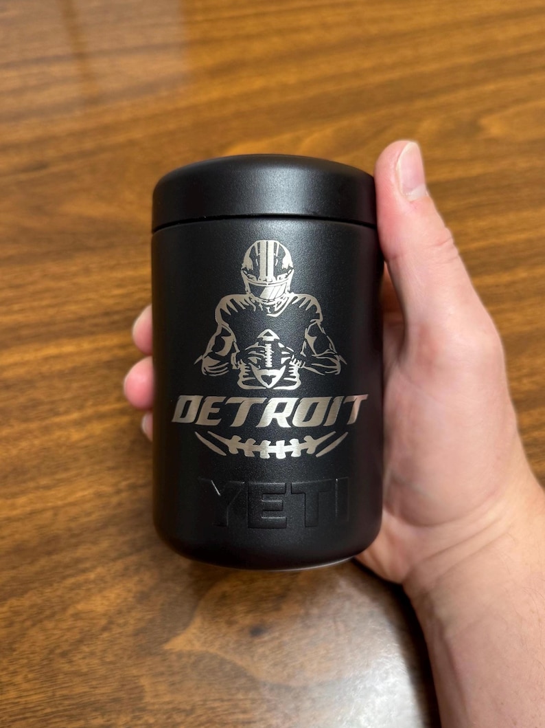 K&ouml;nnte beinhalten: Schwarzer isolierter Becher mit einer silbernen Grafik eines Footballspielers, der einen Football h&auml;lt, dem Wort "DETROIT" und der Marke "YETI". Der Becher ist zylindrisch mit einem schwarzen Deckel.