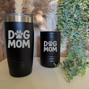 Può includere: Due bicchieri isolati neri con la scritta "DOG MOM" in bianco. Uno è un'alta tazza da viaggio e l'altro è un più piccolo refrigeratore per lattine. Entrambi hanno un design a impronta di zampa. I bicchieri sono su una superficie beige.