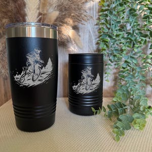 Puede incluir: Juego de vaso y enfriador de latas negros con un diseño de motociclista. El vaso tiene un borde plateado y una tapa transparente. El diseño está grabado en blanco, mostrando a un piloto en una motocicleta en un entorno forestal.