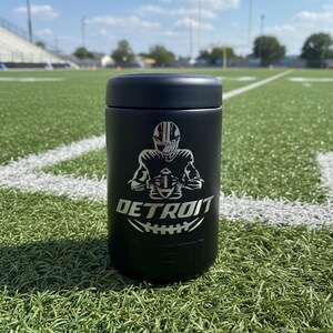 K&ouml;nnte beinhalten: Schwarzer isolierter Becher mit einer silbernen Grafik eines Footballspielers und dem Wort "DETROIT" in Silber. Der Becher ist zylindrisch und hat einen schwarzen Deckel. Der Hintergrund ist ein gr&uuml;nes Footballfeld unter blauem Himmel.