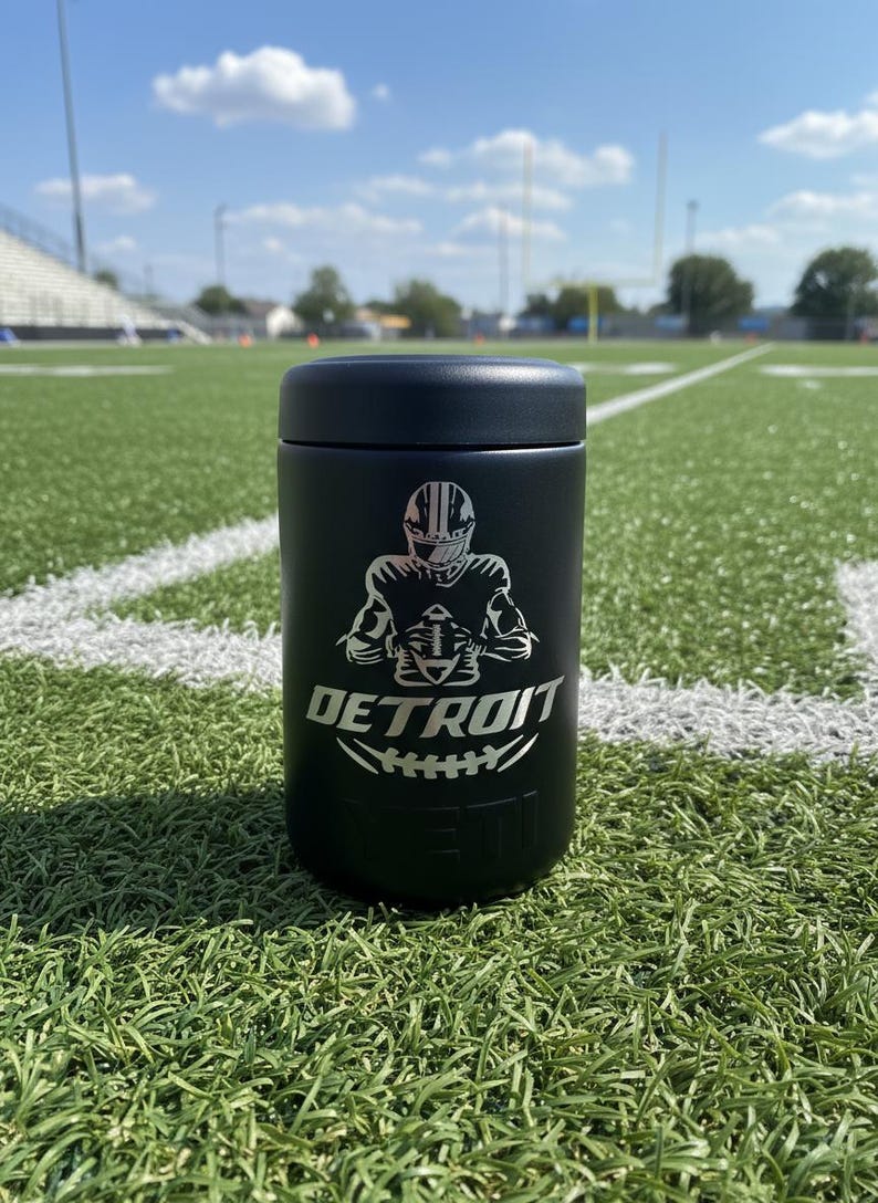 K&ouml;nnte beinhalten: Schwarzer isolierter Becher mit einer silbernen Grafik eines Footballspielers und dem Wort "DETROIT" in Silber. Der Becher ist zylindrisch und hat einen schwarzen Deckel. Der Hintergrund ist ein gr&uuml;nes Footballfeld unter blauem Himmel.