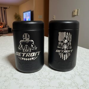 K&ouml;nnte beinhalten: Zwei schwarze isolierte Becher mit silbernen Grafiken. Einer zeigt einen Footballspieler und das Wort "DETROIT" &uuml;ber einem Football. Der andere hat einen Footballspieler mit einem amerikanischen Flaggen-Design. Beide haben den Markennamen "YETI".