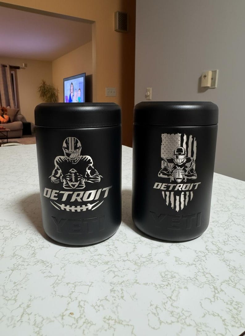 K&ouml;nnte beinhalten: Zwei schwarze isolierte Becher mit silbernen Grafiken. Einer zeigt einen Footballspieler und das Wort "DETROIT" &uuml;ber einem Football. Der andere hat einen Footballspieler mit einem amerikanischen Flaggen-Design. Beide haben den Markennamen "YETI".
