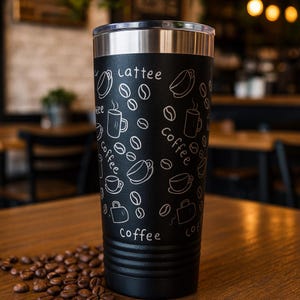 Può includere: Tazza da viaggio nera con bordo argentato, con illustrazioni bianche di chicchi di caffè, tazze e le parole "lattee" e "coffee". La tazza è su un tavolo di legno con chicchi di caffè sparsi.