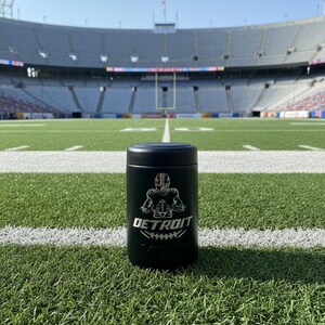 K&ouml;nnte beinhalten: Ein schwarzer Becher mit Football-Motiv, mit einer silbernen Grafik eines Footballspielers und dem Wort "DETROIT". Der Becher steht auf einem gr&uuml;nen Footballfeld mit wei&szlig;en Linien, mit einem Stadion im Hintergrund.