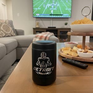 K&ouml;nnte beinhalten: Schwarzer Becher mit blauem Deckel, mit einer Grafik eines Footballspielers aus Detroit und dem Wort "DETROIT". Der Becher steht auf einem Couchtisch aus Holz, im Hintergrund l&auml;uft ein Footballspiel im Fernsehen.