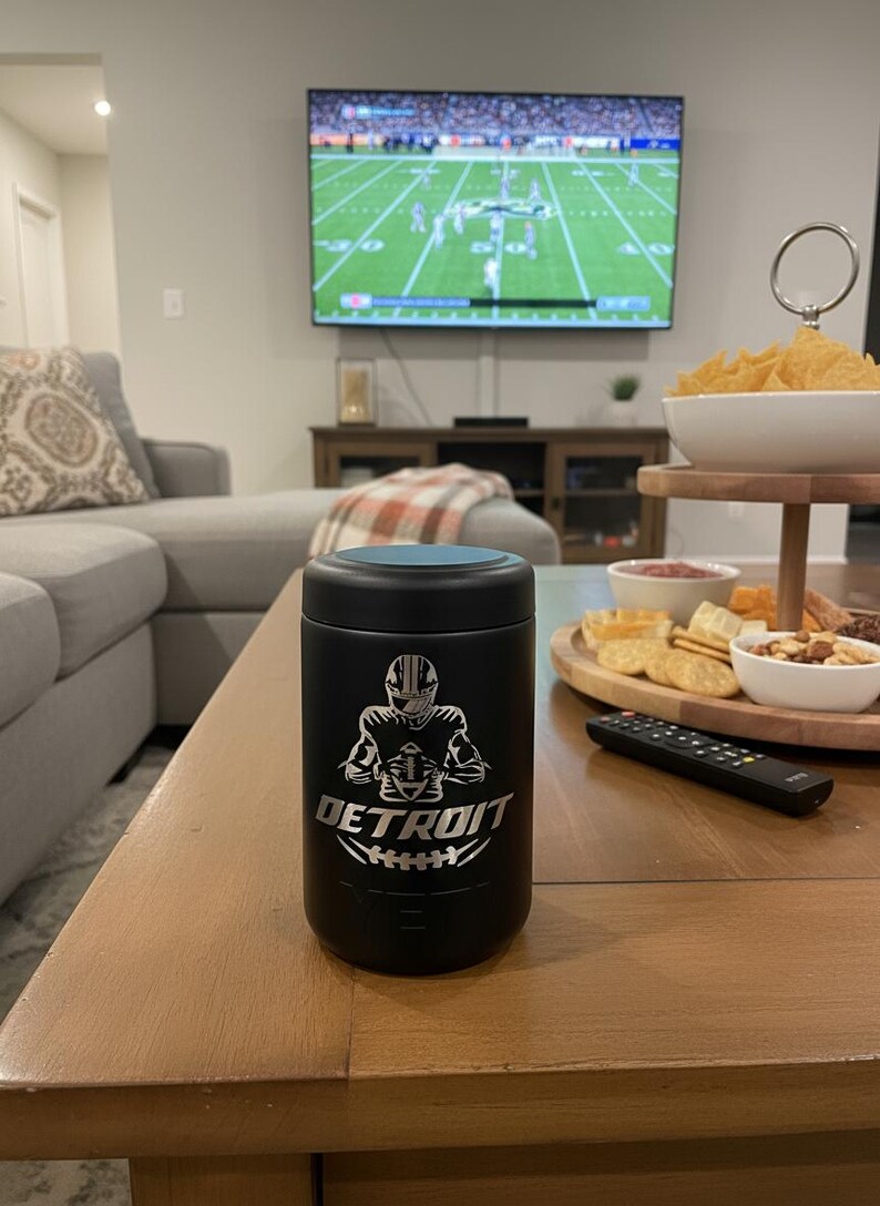 K&ouml;nnte beinhalten: Schwarzer Becher mit blauem Deckel, mit einer Grafik eines Footballspielers aus Detroit und dem Wort "DETROIT". Der Becher steht auf einem Couchtisch aus Holz, im Hintergrund l&auml;uft ein Footballspiel im Fernsehen.