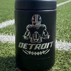K&ouml;nnte beinhalten: Schwarzer isolierter Becher mit einer silbernen Grafik eines Footballspielers und dem Wort "DETROIT" mit einem Football-Design. Der Becher ist zylindrisch und hat einen schwarzen Deckel. Der Hintergrund ist ein gr&uuml;nes Feld.