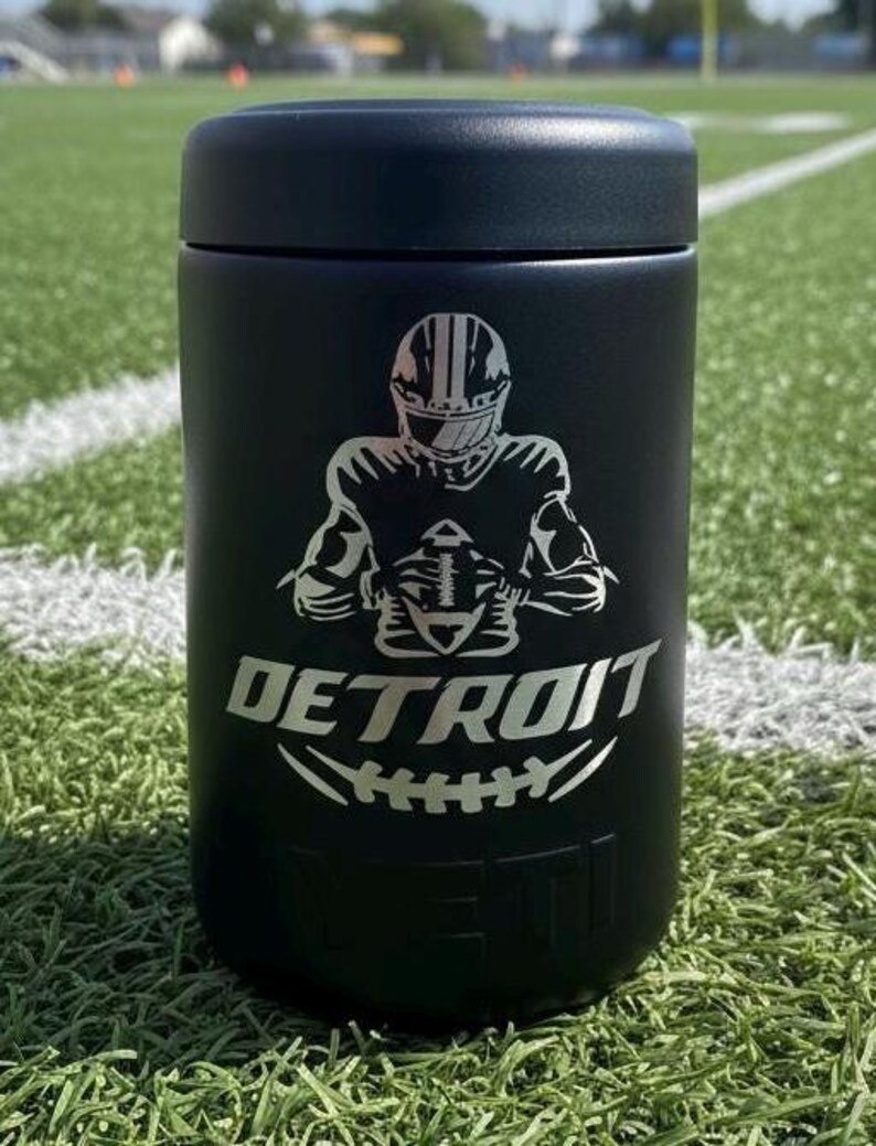 K&ouml;nnte beinhalten: Schwarzer isolierter Becher mit einer silbernen Grafik eines Footballspielers und dem Wort "DETROIT" mit einem Football-Design. Der Becher ist zylindrisch und hat einen schwarzen Deckel. Der Hintergrund ist ein gr&uuml;nes Feld.