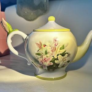 May include: A white ceramic teapot featuring a yellow lid and base trim. The body displays a floral design with white and pink lilies and a small hummingbird. A three-dimensional hummingbird figure is attached to the handle of the teapot.