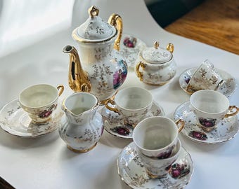 May include: A vintage porcelain tea set with a white and gold floral design. The set includes a teapot, sugar bowl, creamer, and several teacups with saucers. The teapot and teacups have gold handles and accents.