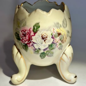 May include: Cream-colored, egg-shaped ceramic vase with three curved feet. The vase features hand-painted roses in shades of red, white, and yellow, with green leaves. Gold trim accents the rim and feet, adding a touch of elegance to this decorative piece.