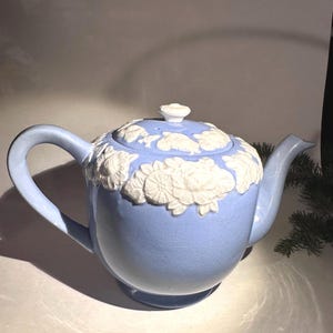 May include: A light blue ceramic teapot with a white floral design around the lid. The teapot has a curved handle and spout. The lid has a small white knob. The teapot is a decorative item.