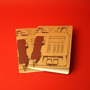 Könnte beinhalten: Zwei beigefarbene Notizbücher mit einer dunkelbraunen Strichzeichnung eines alten öffentlichen Telefons auf dem Einband. Die Illustration zeigt einen Hörer, ein Tastenfeld und den deutschen Text "Wunschnummer" über dem Tastenfeld.