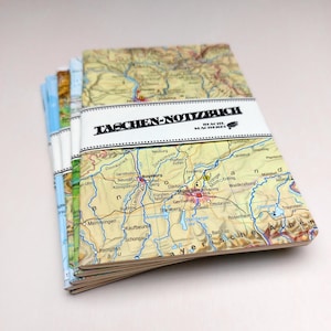 Könnte beinhalten: Ein Stapel Notizbücher mit Kartenmotiven. Die Notizbücher sind mit einem weißen Band gebunden, auf dem "TASCHEN-NOTIZBUCH" steht. Die Karten zeigen verschiedene Städte und geografische Details, mit Schwerpunkt auf der Region München.