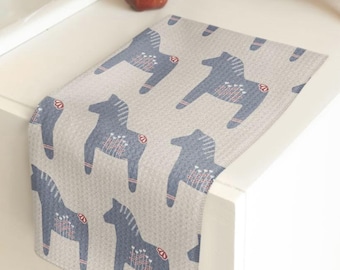 May include: A rectangular kitchen towel with a light gray waffle-textured background featuring a repeating pattern of blue stylized horse figures. Each horse has white and red decorative line details on its body and mane.