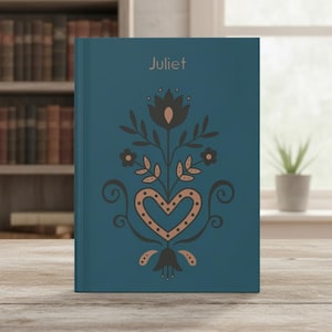 May include: A teal hardback book featuring the name Juliet printed in light brown at the top. The cover displays a symmetrical floral and heart-shaped graphic design in dark grey and light brown, centred vertically on the front cover.