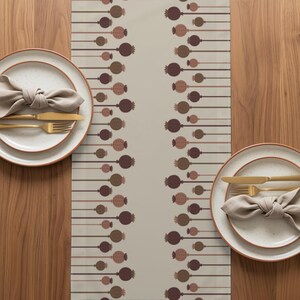 May include: A table runner with a beige background and a repeating vertical pattern of brown and tan poppy seed pods on thin stems. Two place settings feature white speckled plates, beige cloth napkins, and gold-toned flatware.