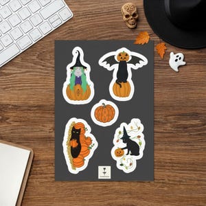 Peut inclure: Une planche d'autocollants présentant cinq illustrations sur le thème d'Halloween sur un fond gris foncé. Les motifs incluent un personnage aux cheveux verts avec un chapeau de sorcière dans une citrouille, un chat noir ailé sur une citrouille, une citrouille, un chat noir avec des citrouilles et un chat noir avec un chapeau de sorcière et des vignes.