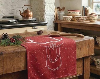 May include: A red rectangular fabric runner featuring a white line-art illustration of a reindeer head with a snowflake on its forehead and the text Merry Christmas, accented by a subtle white star pattern across the surface.