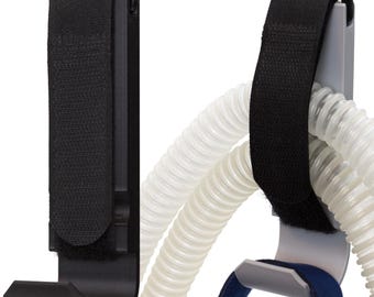 May include: Two black CPAP hose holders with a hook design. The holders have a black fabric strap with a hook and loop closure. White, flexible tubing is held by the holders. A blue strap is also visible.