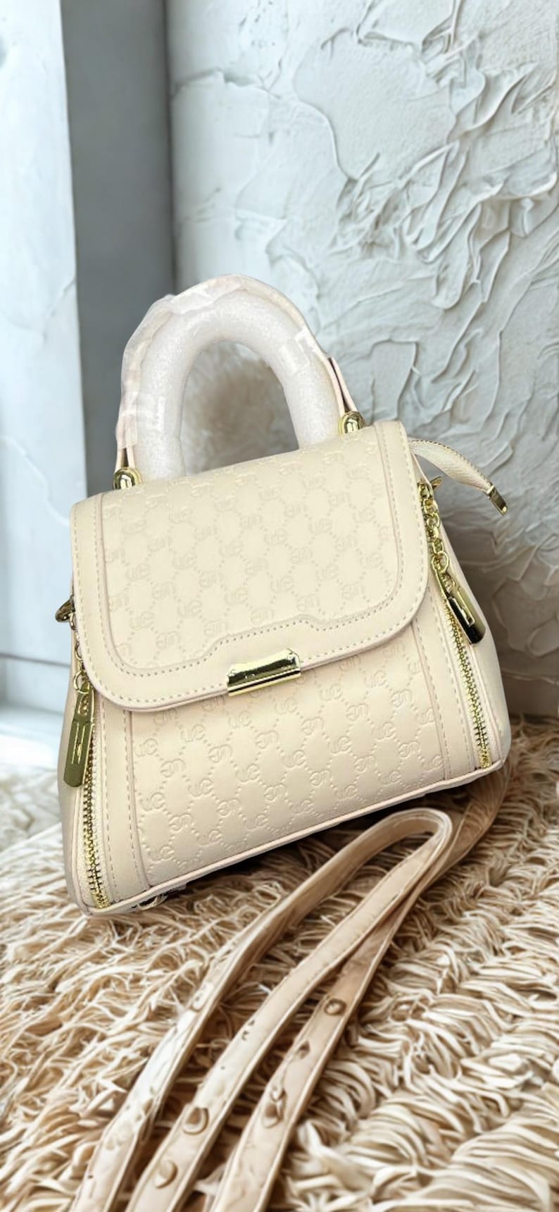 May include: Cream-coloured handbag with a top handle and a detachable shoulder strap. The bag features a repeating pattern and gold-toned hardware, including zips and a clasp. The bag is sitting on a textured surface.
