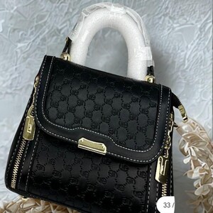 May include: Black handbag with a repeating pattern, white stitching, and gold-tone hardware. Features a top handle and a detachable shoulder strap. The bag has a zipper closure and a small charm. The number "33" is visible on the side.