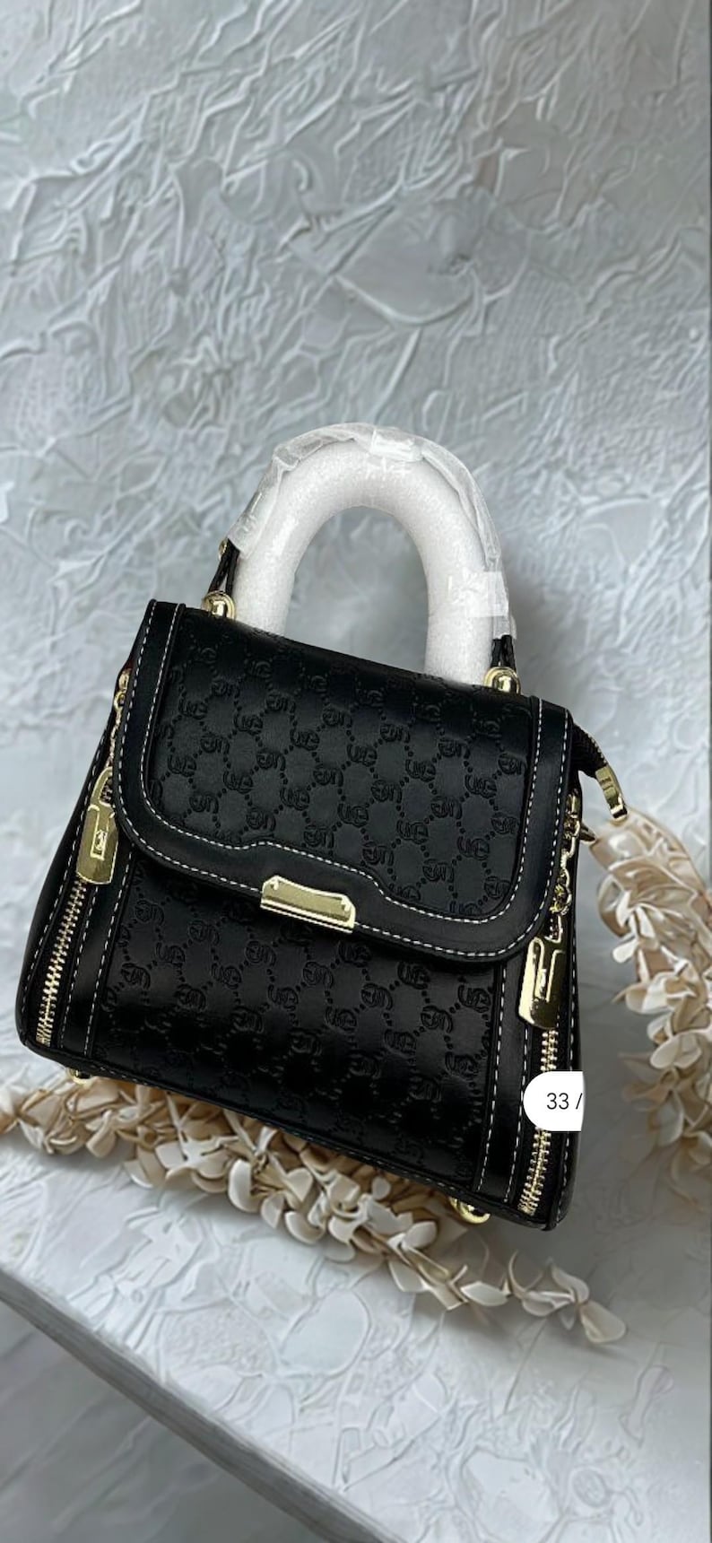 May include: Black handbag with a repeating pattern, white stitching, and gold-tone hardware. Features a top handle and a detachable shoulder strap. The bag has a zipper closure and a small charm. The number "33" is visible on the side.