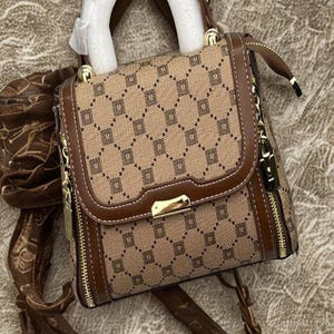 May include: A brown and tan handbag featuring a repeating "M" pattern. It has a top handle and a long strap. Gold-tone zips and hardware provide detail. The bag rests on a textured surface. The bag's dimensions are approximately 20 cm x 20 cm.