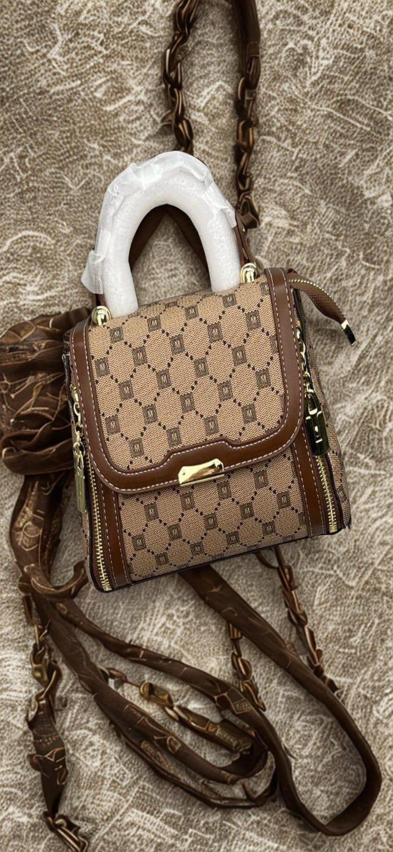 May include: A brown and tan handbag featuring a repeating "M" pattern. It has a top handle and a long strap. Gold-tone zips and hardware provide detail. The bag rests on a textured surface. The bag's dimensions are approximately 20 cm x 20 cm.