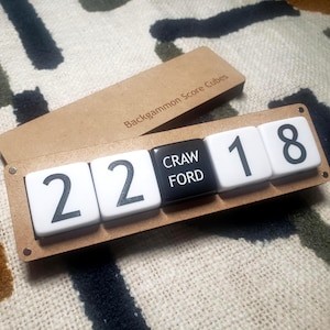 Puede incluir: Cubos de puntuación de backgammon con el texto "Backgammon Score Cubes" en la tapa. Los cubos muestran los números 22, CRAWFORD, 18. Los cubos son blancos con números y letras negras, y el cubo CRAWFORD es negro con letras blancas.