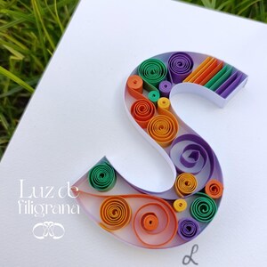 Può includere: Un'opera d'arte di quilling di carta fatta a mano con la lettera "S". La lettera è riempita con spirali di carta colorate nelle tonalità di arancione, verde, viola e giallo. L'opera è su sfondo bianco con il testo "Luz de filigrana".