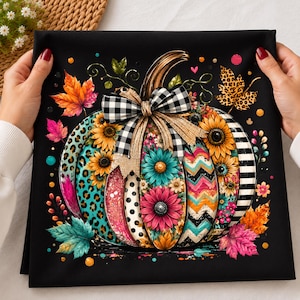 May include: A black fabric panel featuring a central pumpkin illustration with segments decorated in leopard print, black and white polka dots, teal and pink flowers, and chevron patterns. A black and white buffalo plaid bow sits on top.