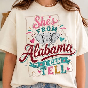May include: A cream-colored t-shirt featuring a graphic of the state of Alabama with an elephant illustration. The text reads "She's FROM ALABAMA I CAN TELL" in pink, teal, and dark red. The design includes small hearts and stars in a distressed style.