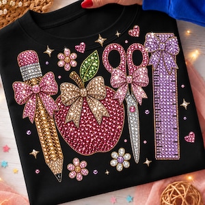 May include: A black t-shirt featuring a graphic design composed of sparkling rhinestones. The design includes a pencil, an apple, scissors, and a ruler, all adorned with decorative bows, surrounded by small flower and star accents.