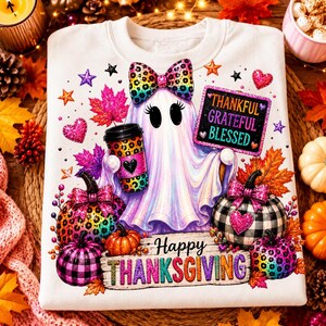 Op de afbeelding: Een wit sweatshirt met een spookafbeelding met een kleurrijke strik met luipaardprint, die een bijpassende koffiebeker vasthoudt. Het ontwerp bevat pompoenen, herfstbladeren, hartjes, sterren en de tekst Thankful Grateful Blessed.