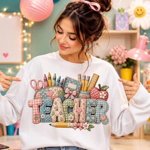 May include: A white long-sleeve sweatshirt featuring a graphic print of the word TEACHER in a quilted fabric pattern. The letters are decorated with illustrations of scissors, crayons, a notebook, pencils, a ruler, and a red apple with a flower.