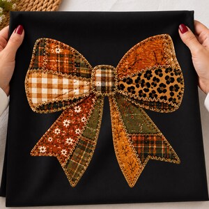 May include: A black fabric square featuring a large, central bow appliqué. The bow is constructed from various patchwork fabric segments in shades of orange, brown, green, and cream, showcasing plaid, leopard print, and floral patterns with gold stitching.