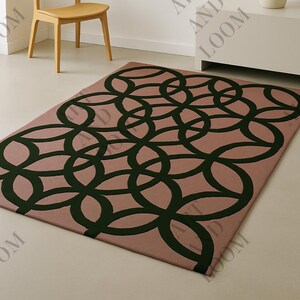May include: A rectangular rug featuring a geometric design of dark green interlocking circles on a dusty rose backdrop. The rug is in a room with a wooden chair, a white cabinet, and a white vase.