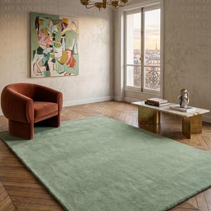 May include: A large, rectangular, sage green rug dominates a room with hardwood floors. An abstract painting hangs on the wall, and a rust-colored armchair sits near the rug. A brass chandelier hangs from the ceiling.