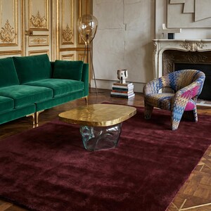 May include: A luxurious living room setting featuring a deep burgundy rug, a green velvet sofa, and a patterned armchair. A unique coffee table with a gold top and glass base sits atop the rug. The room has ornate gold wall paneling and a fireplace.
