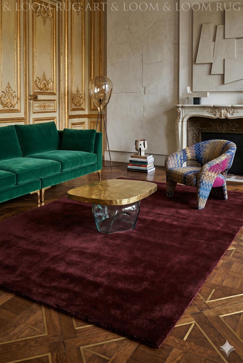May include: A luxurious living room setting featuring a deep burgundy rug, a green velvet sofa, and a patterned armchair. A unique coffee table with a gold top and glass base sits atop the rug. The room has ornate gold wall paneling and a fireplace.