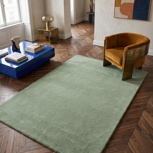 May include: A large, rectangular sage green rug in a living room setting. The rug is placed on a dark wood parquet floor. An orange armchair and a blue coffee table are also visible. The room has a modern, minimalist aesthetic.