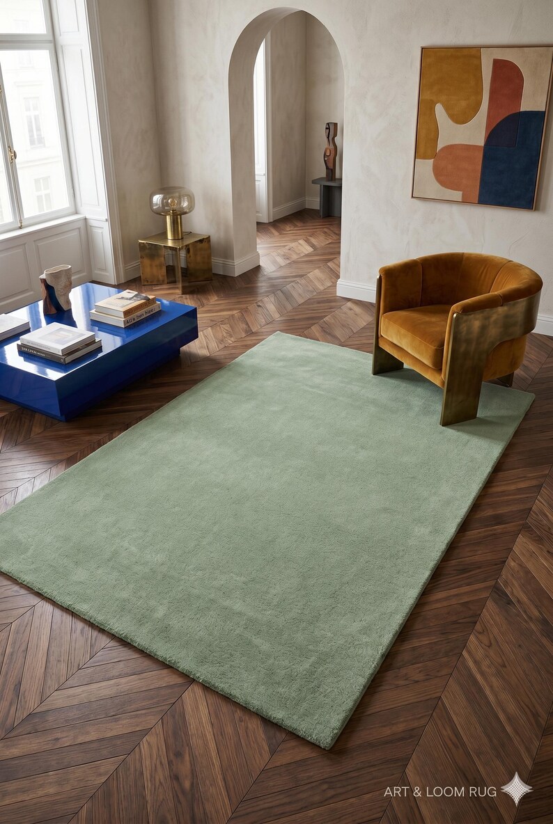 May include: A large, rectangular sage green rug in a living room setting. The rug is placed on a dark wood parquet floor. An orange armchair and a blue coffee table are also visible. The room has a modern, minimalist aesthetic.