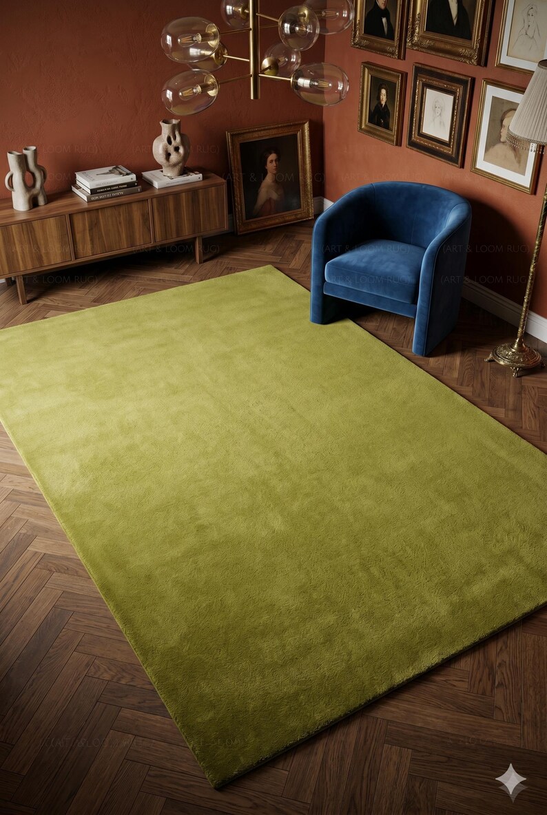 May include: A large, rectangular, olive-green area rug. The rug is the focal point of a room with a wooden floor, a blue armchair, and a wooden cabinet. The walls are painted a warm, reddish-brown color.