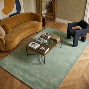 May include: A sage green rectangular rug in a living room setting. The rug is placed on a wooden floor with a herringbone pattern. A gold-toned coffee table with books and a glass bowl sits on the rug. A mustard-colored sofa and a dark blue chair are also in the room.