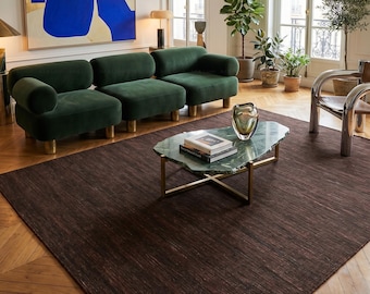 Op de afbeelding: Een woonkamer met een donkerbruin tapijt, smaragdgroene fluwelen banken met gouden poten en een marmeren salontafel. Een groot abstract schilderij met blauwe en gouden tinten hangt aan de muur. De kamer heeft houten vloeren en grote ramen.