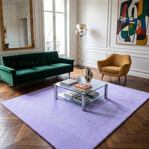 May include: A large, rectangular, light purple rug in a living room setting. The rug is placed on a herringbone patterned hardwood floor. A green velvet sofa, a yellow armchair, and a glass coffee table are also visible.